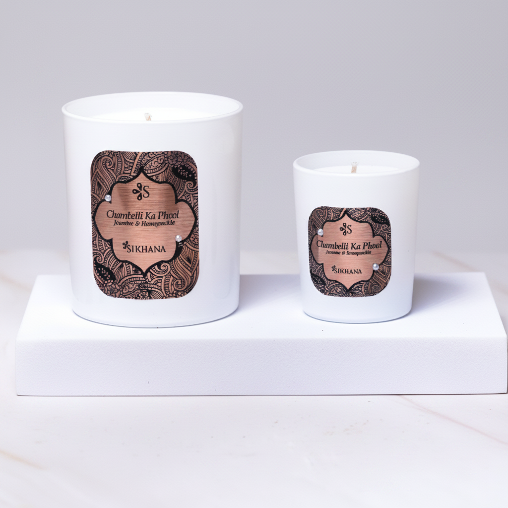 Two white candles with decorative labels on a light surface with flowers and leaves.