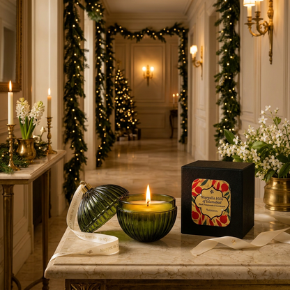A lit green ribbed candle in a dark green glass holder sits on a marble table beside a black gift box with a floral label, set in a decorated holiday hallway with garlands and warm wall lights.