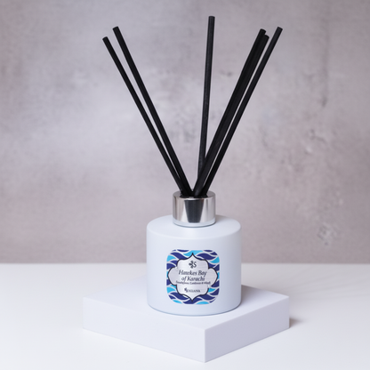 Diffuser with reeds and a decorative label on a white surface with green leaves in the background