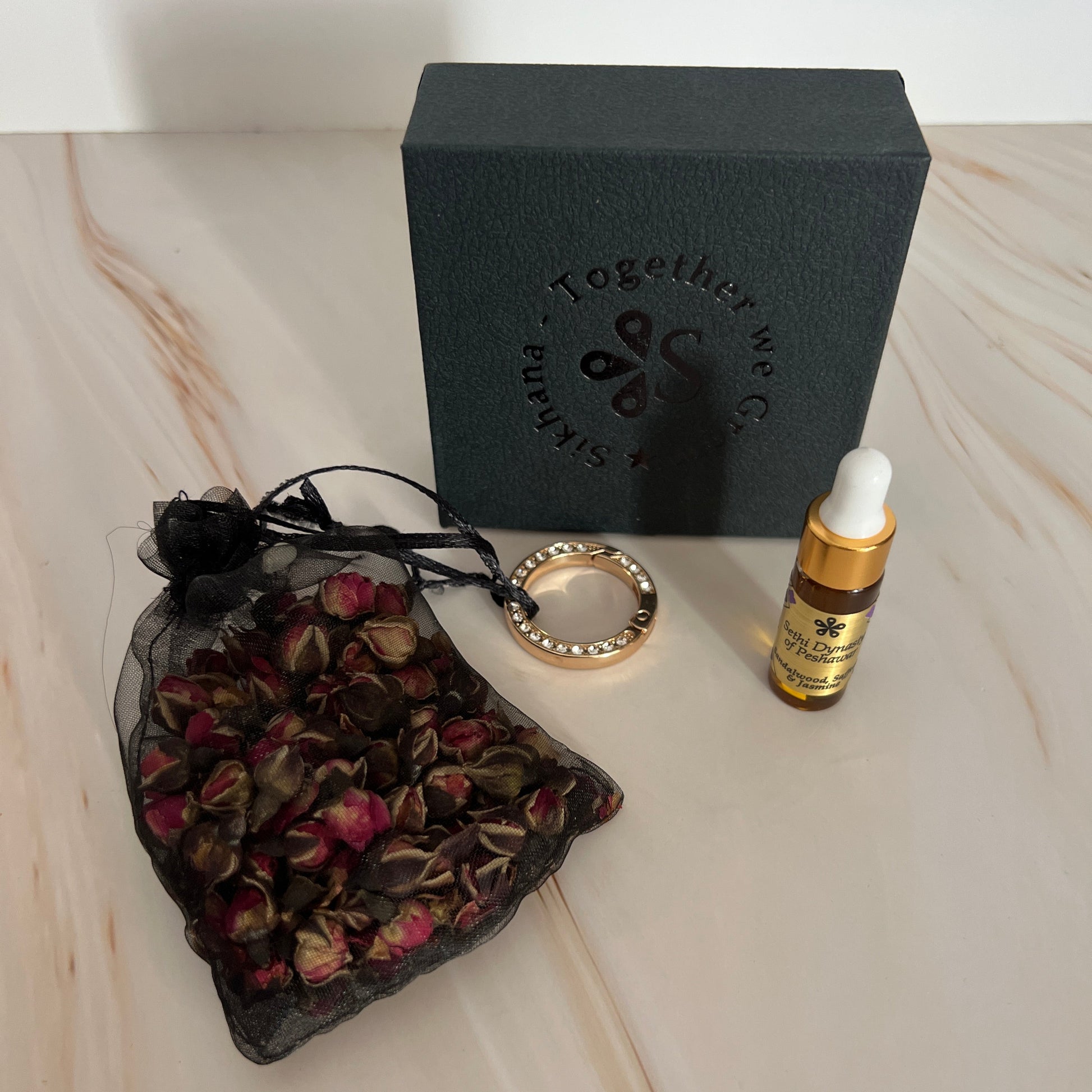 Black box with a logo, gold pen, ring, and black bag with dried flowers on a marble surface.