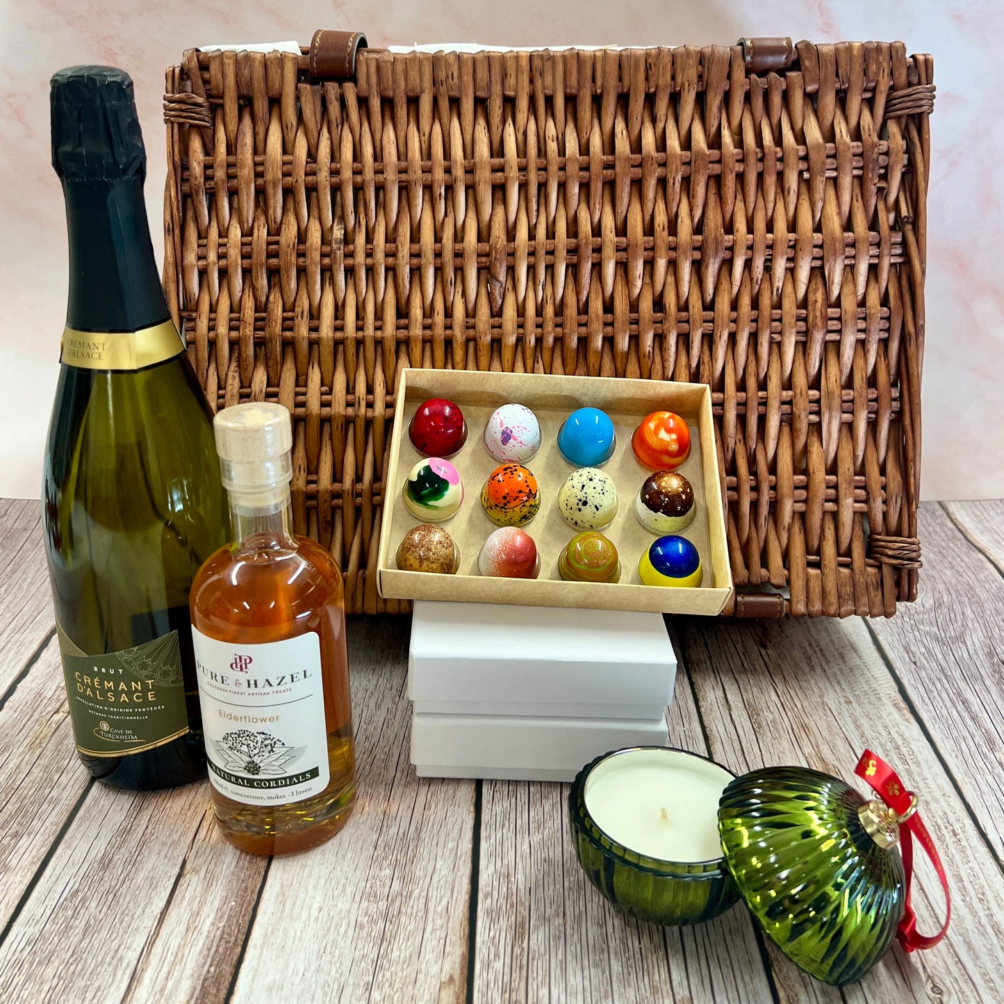 Wicker hamper with bottles, a box of chocolates, and decorative items on a wooden surface.