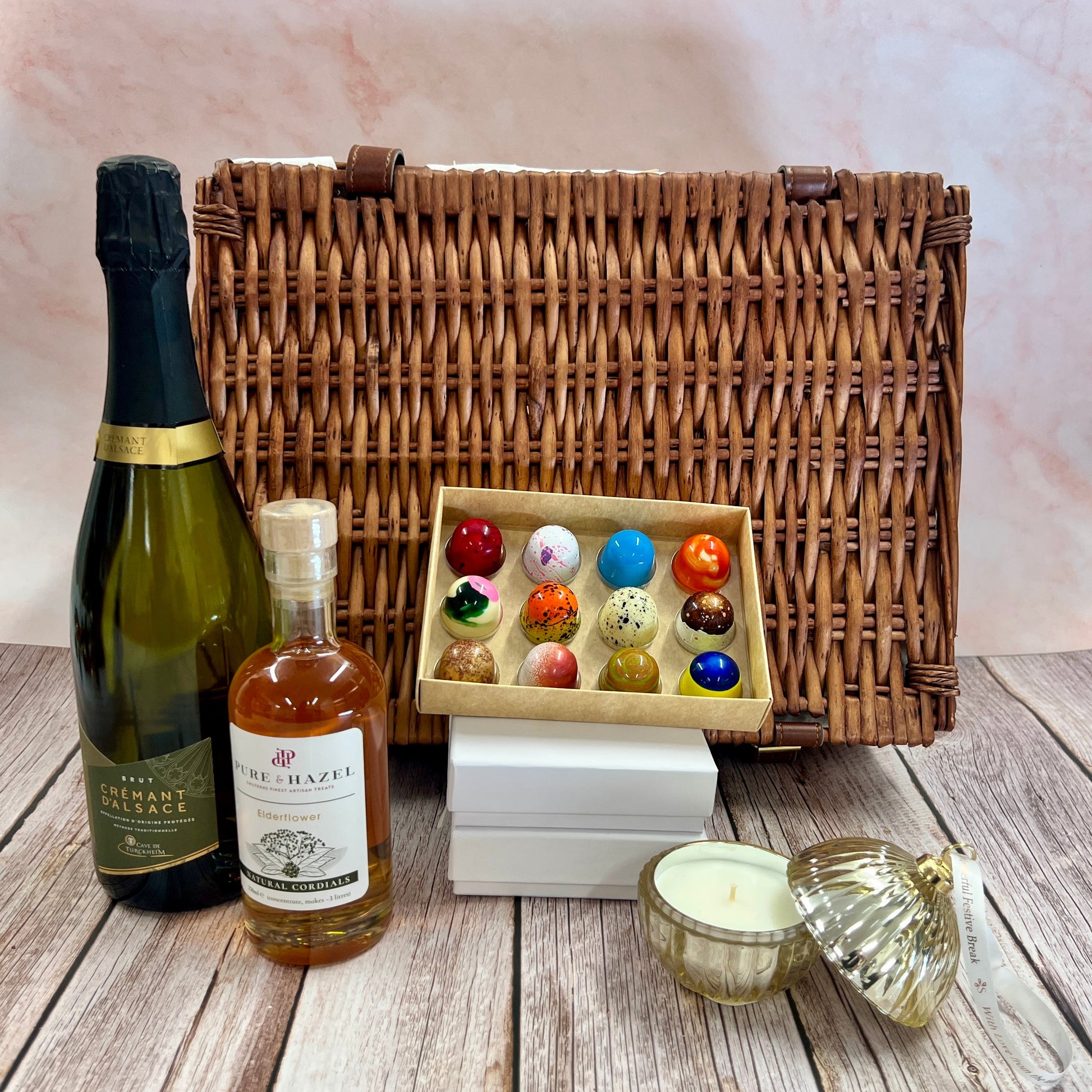 Wicker picnic basket with bottles, a box of colorful candies, and a candle on a wooden surface.