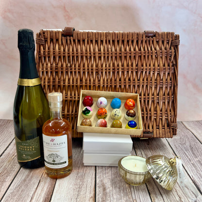 Wicker picnic basket with bottles, a box of colorful candies, and a candle on a wooden surface.