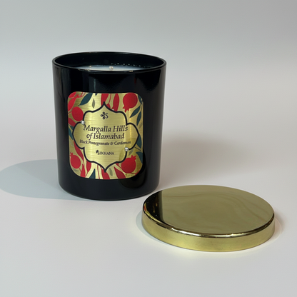 A black candle with a label that reads 'MARGALLA HILLS OF ISLAMABAD', placed on a marble surface, with a gold lid on the side.