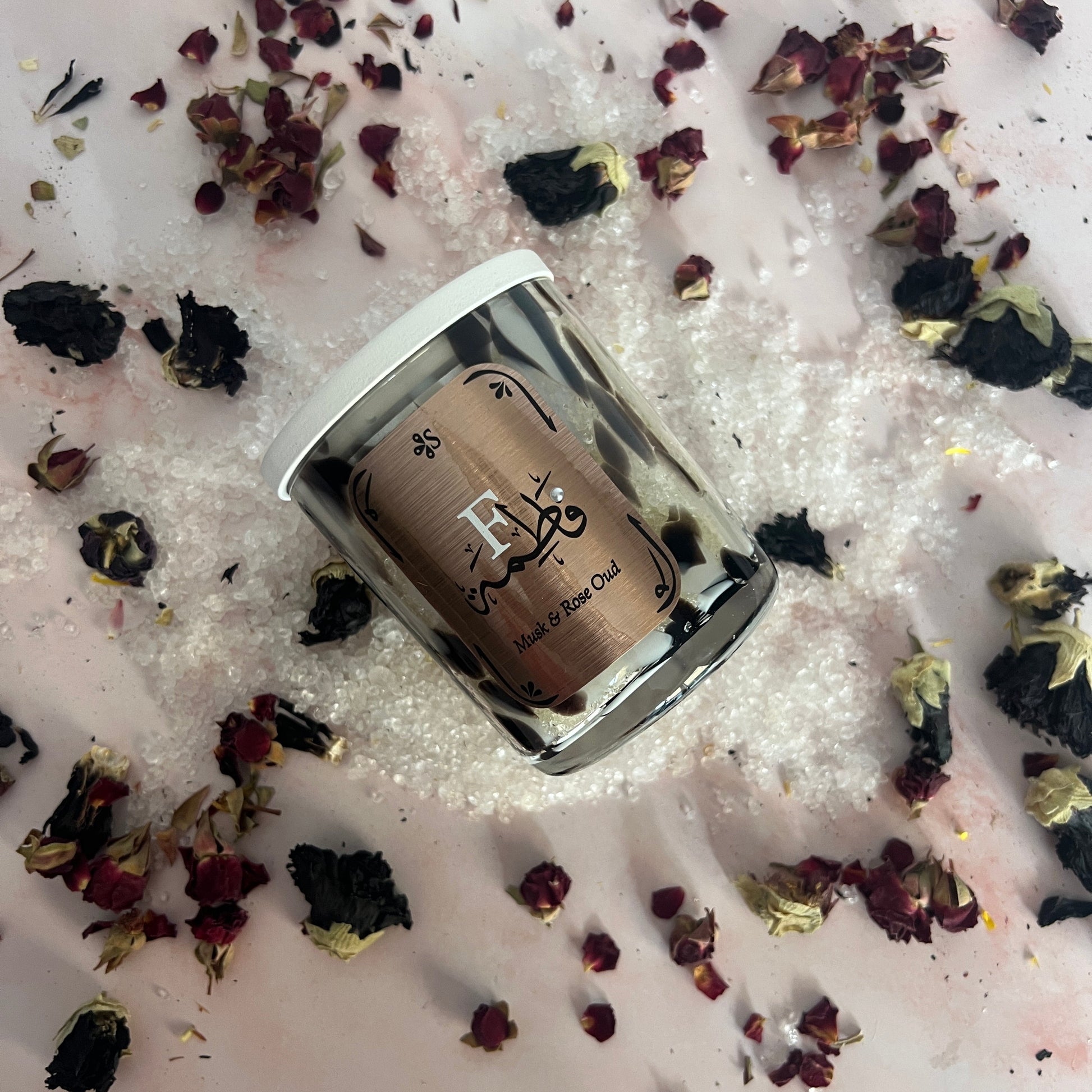 Small container with a decorative label on a bed of pink bath salts and dried flowers.