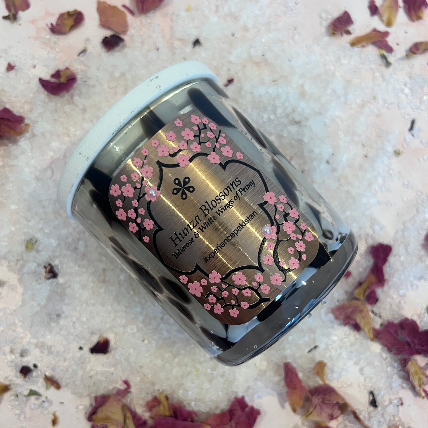 Small cylinder container with floral design on a bed of pink rose petals and white sand.