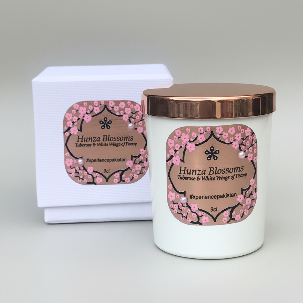 A white candle with a copper lid labeled 'Hunza Blossoms' and pink floral designs, placed next to its luxury rigid black gift box with foil labels and pearl embellishments.