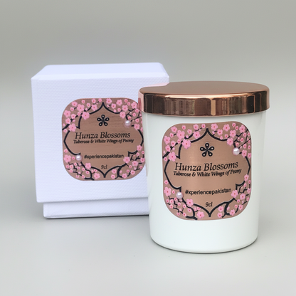 A white candle with a copper lid labeled 'Hunza Blossoms' and pink floral designs, placed next to its luxury rigid black gift box with foil labels and pearl embellishments.