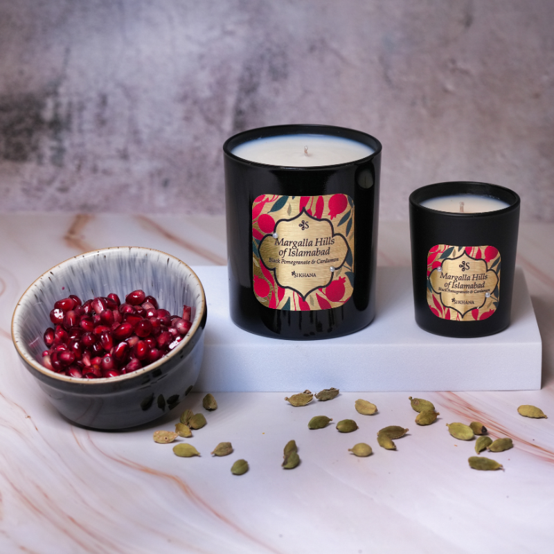 Two black candles with floral labels on a marble surface with a bowl of red seeds and green cardamom pods.